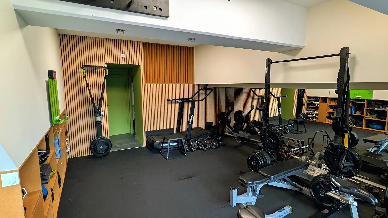 Le studio en journée : mur en tasseaux de bois, porte vert pomme, ski erg, tapis incurvé, kettlebells, rameurs et rack devant le miroir
