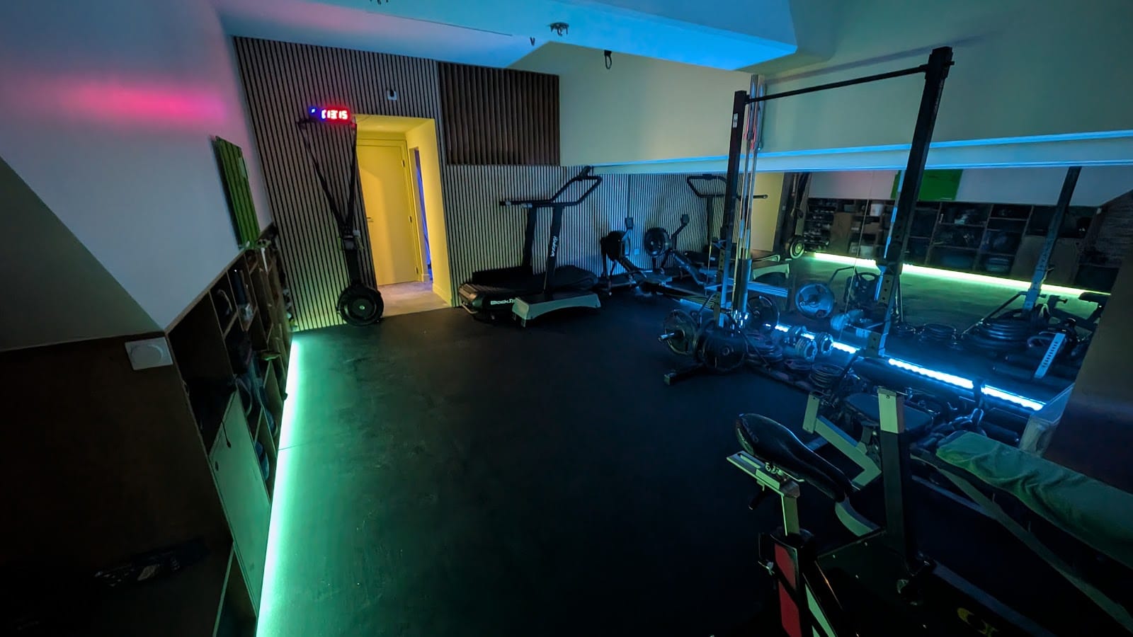 La salle du studio la nuit : LED vertes et cyan, chrono rouge au-dessus de la porte éclairée, rack, rameurs et tapis incurvé, sol noir